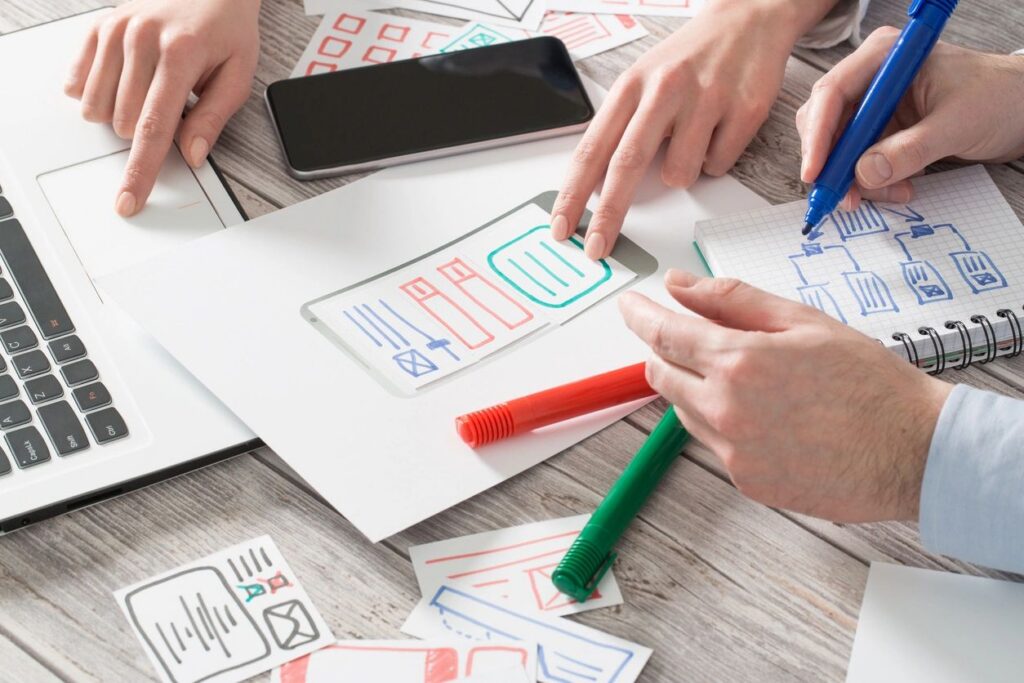 Hände ordnen Ideen auf Papier mit Skizzen und bunten Stiften, daneben ein Smartphone auf dem Schreibtisch. zu viele Tabs im Kopf.